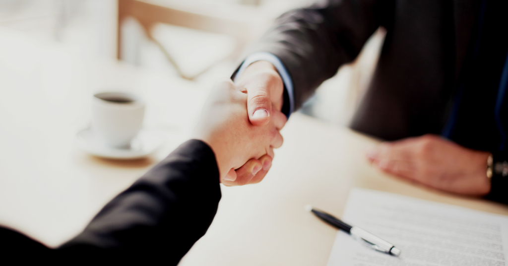Two business professionals shaking hands at a table with a contract and coffee, representing partnership agreements in IT outsourcing.