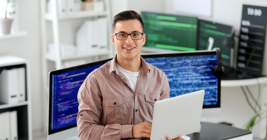A confident software developer holding a laptop in front of multiple monitors with code, representing a productive and optimized development environment.