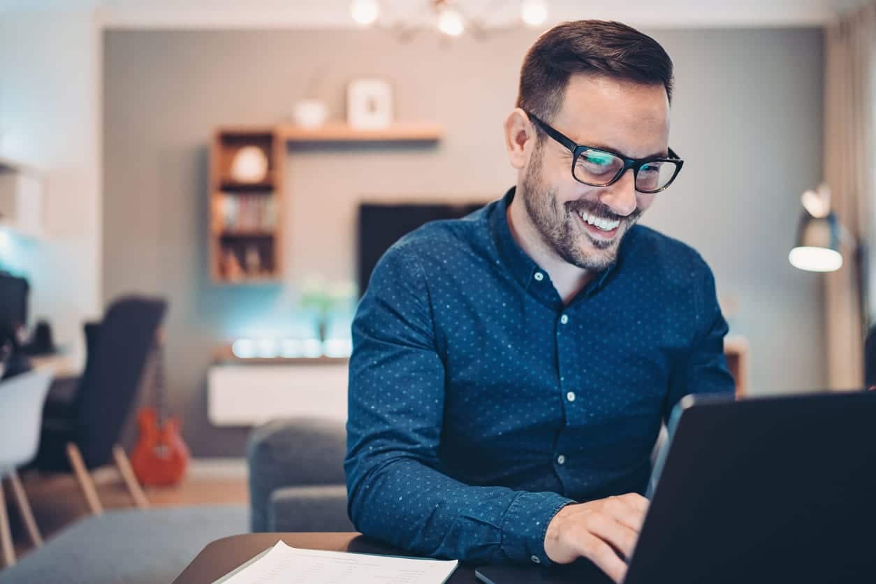 smiling-man-sitting-at-desk-at-home-with-open-laptop-and-living-room-behind-him