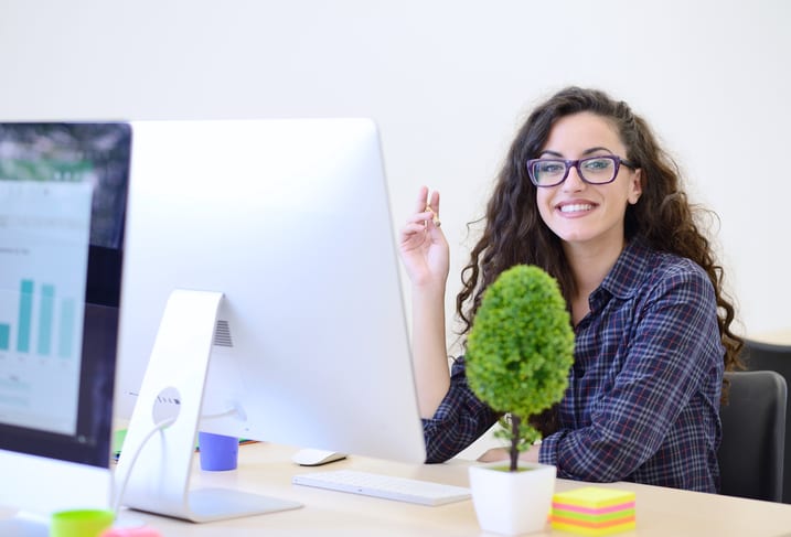 woman-seated-at-desk-with-computer-monitor-and-potted-plant-while-smiling-at-camera