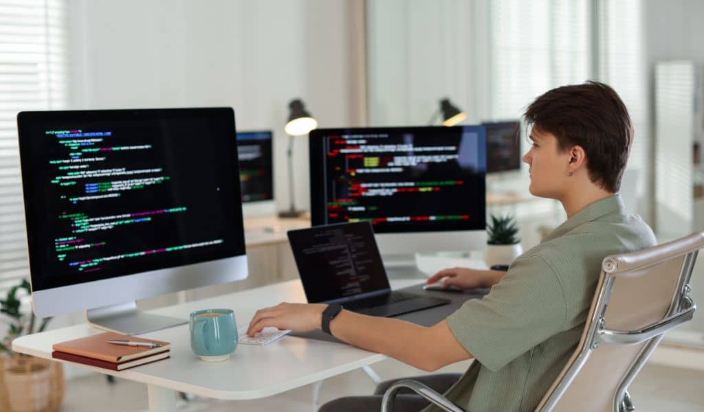 parallelstaff- A person sits at a desk using a laptop with two large monitors displaying colorful lines of code, in a bright office with modern decor and potted plants—a perfect setting for nearshore software development.