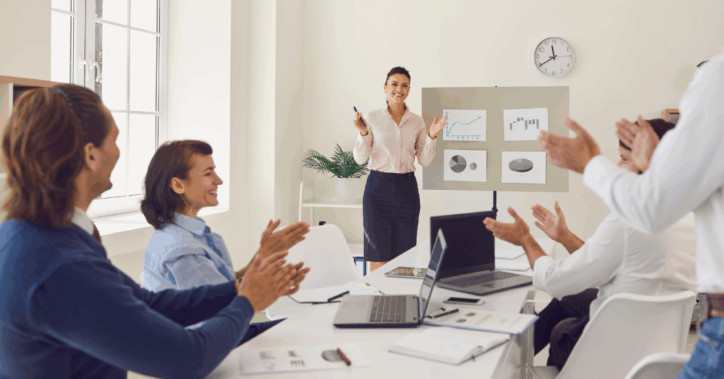 Business team applauding a female presenter in a meeting room with performance charts behind her.