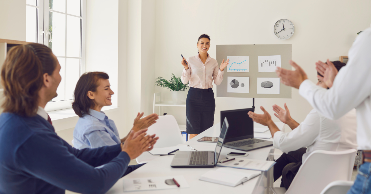 Business team applauding a female presenter in a meeting room with performance charts behind her.