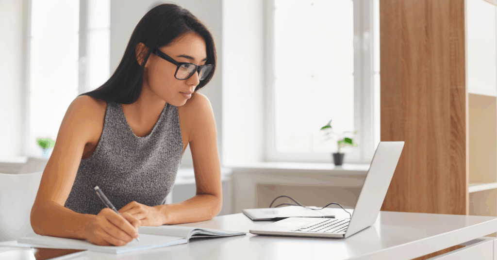 Focused woman writing notes while looking at her laptop, analyzing professional alternatives for hiring skilled developers.