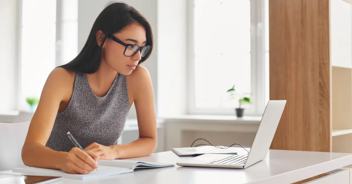 Focused woman writing notes while looking at her laptop, analyzing professional alternatives for hiring skilled developers.
