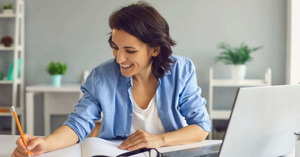 Woman smiling and writing notes while working remotely with a laptop on desk.