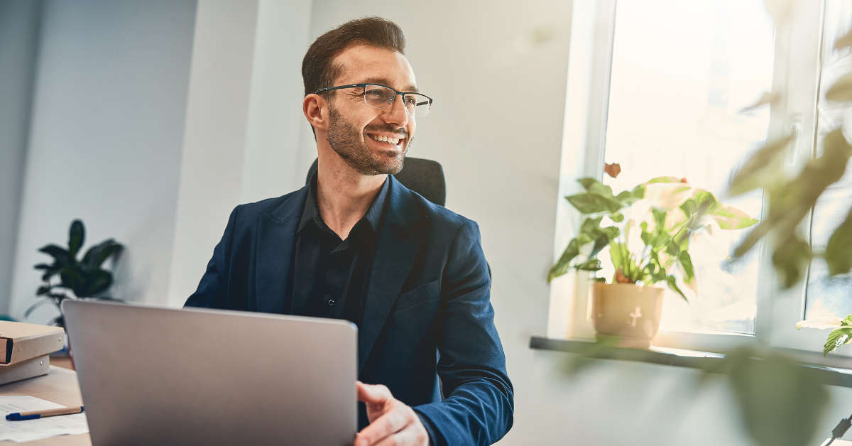 Confident man smiling while working on his laptop in a bright office with plants.