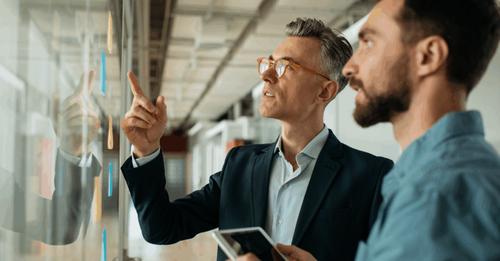 Two professionals pointing at sticky notes on a glass wall, organizing tasks in a project planning session aligned with agile DevOps.