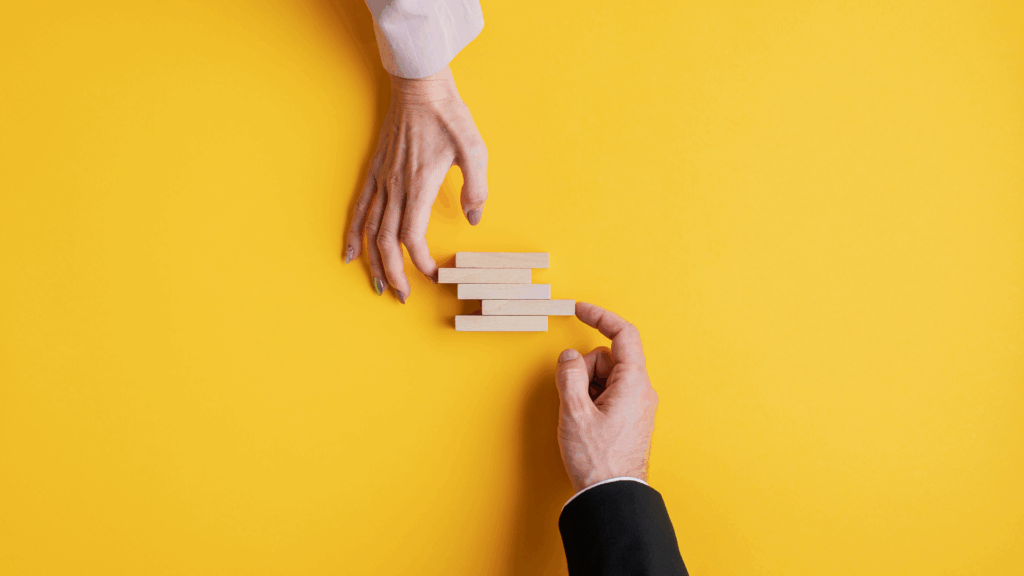 Close-up of two hands from business professionals aligning wooden blocks on a vibrant yellow background, illustrating partnership and structure.
