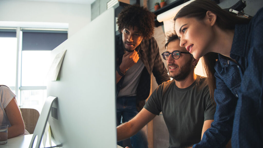 parallelstaff- Three young adults collaborate at a computer in a bright, modern office. Focused on the screen, they embody teamwork—an ideal scene for startups seeking affordable IT staff augmentation services. Large windows and plants add to the vibrant space.