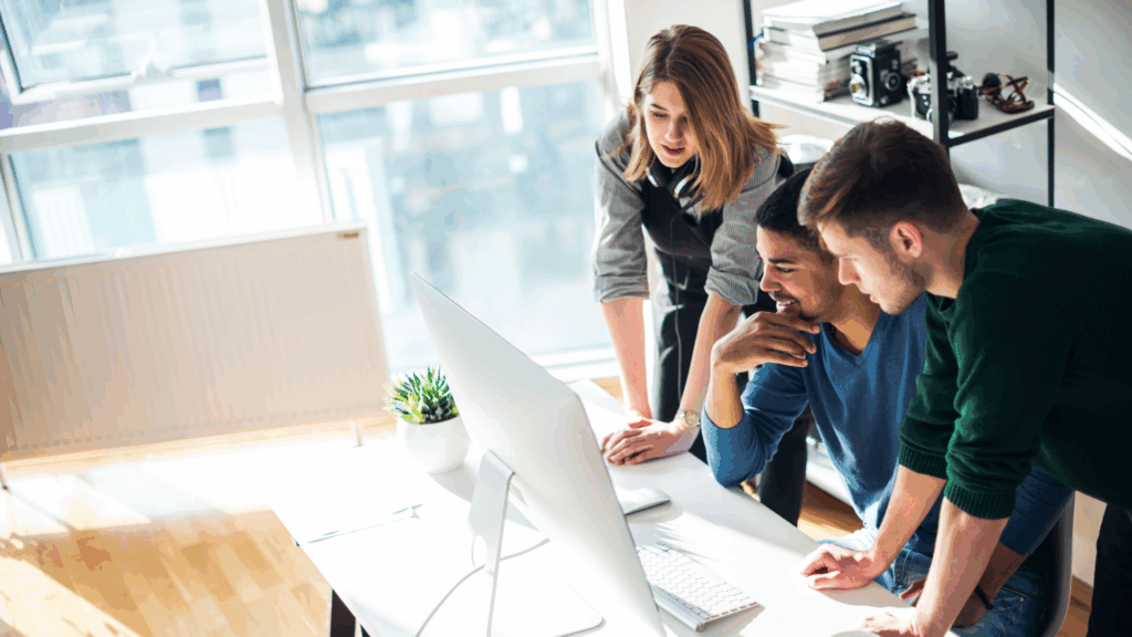 Three team members gathered around a computer in a bright office, collaborating on a project.