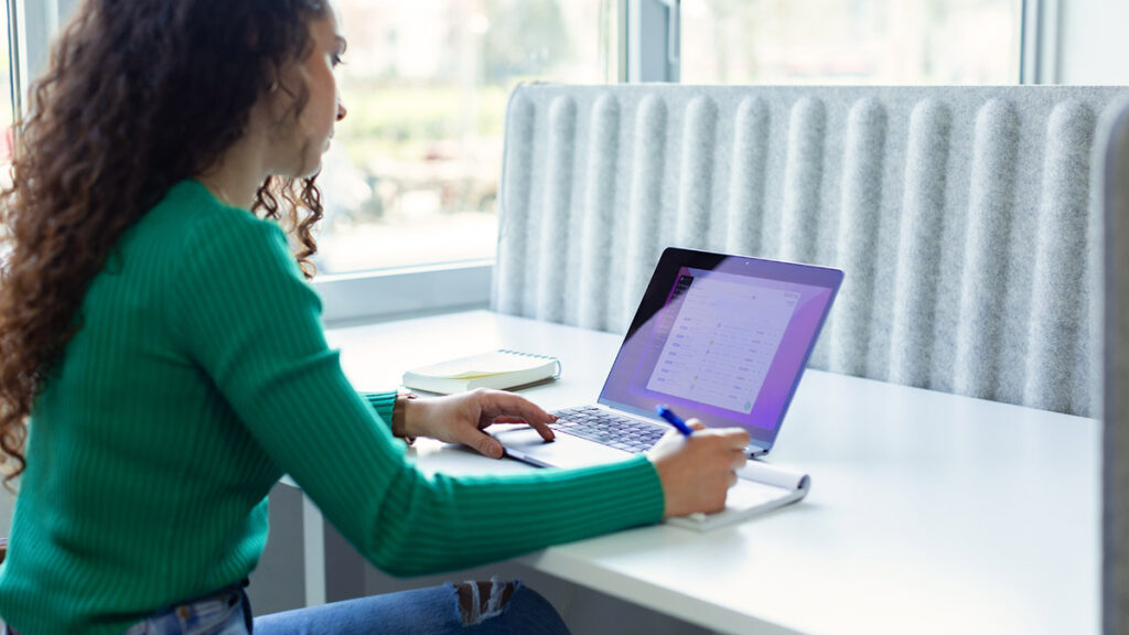parallelstaff- A woman with curly hair, wearing a green sweater, sits at a white desk by a window. She is researching affordable IT staff augmentation services for startups on her laptop while jotting notes in her notebook.