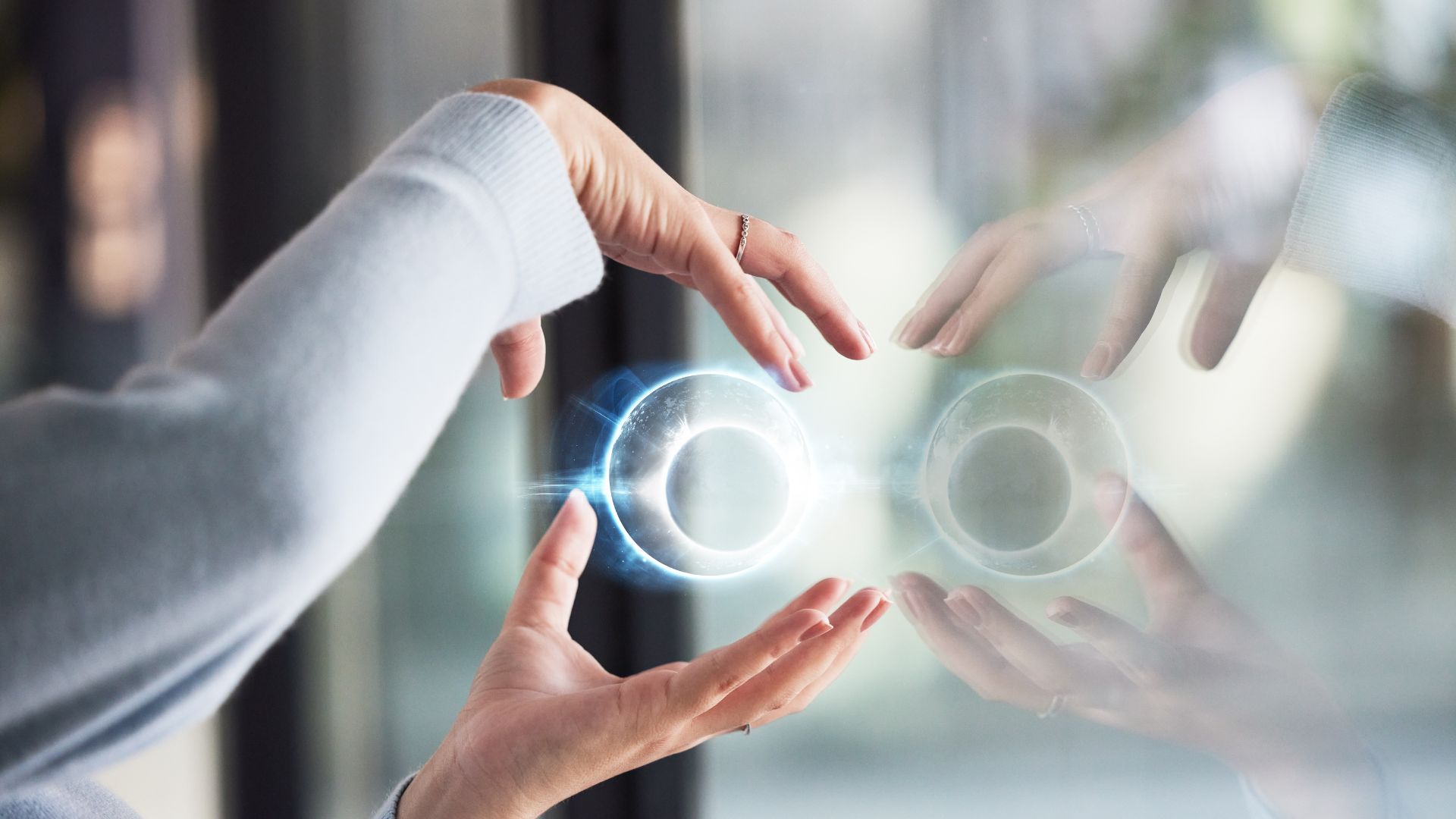 parallelstaff- A person’s hands hold a glowing, futuristic orb near a glass surface, creating a bright reflection. The background is blurred, focusing attention on the orb and hands.