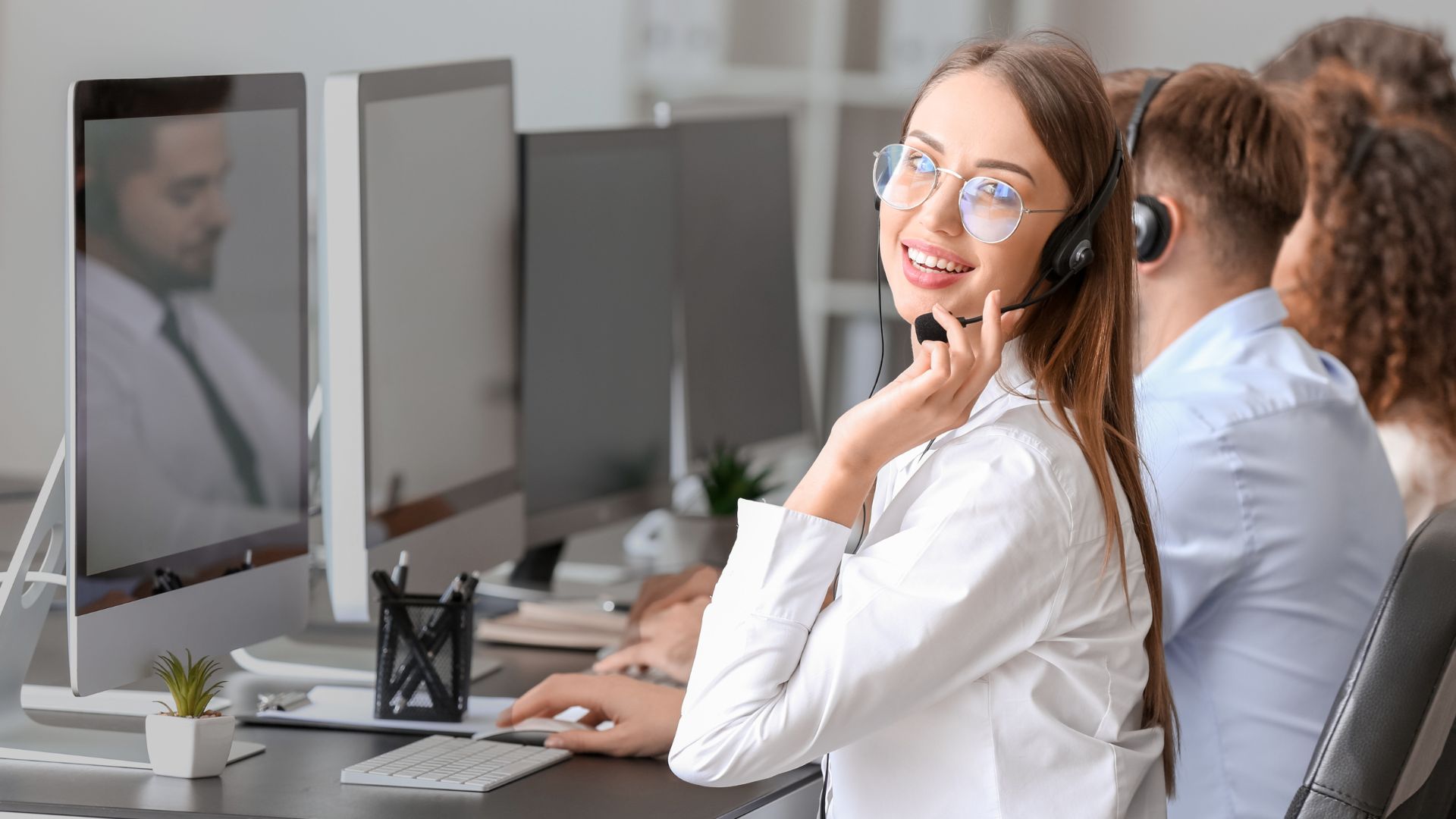 parallelstaff- A woman wearing glasses and a headset smiles at the camera while working at a computer in an office with colleagues at adjacent desks.