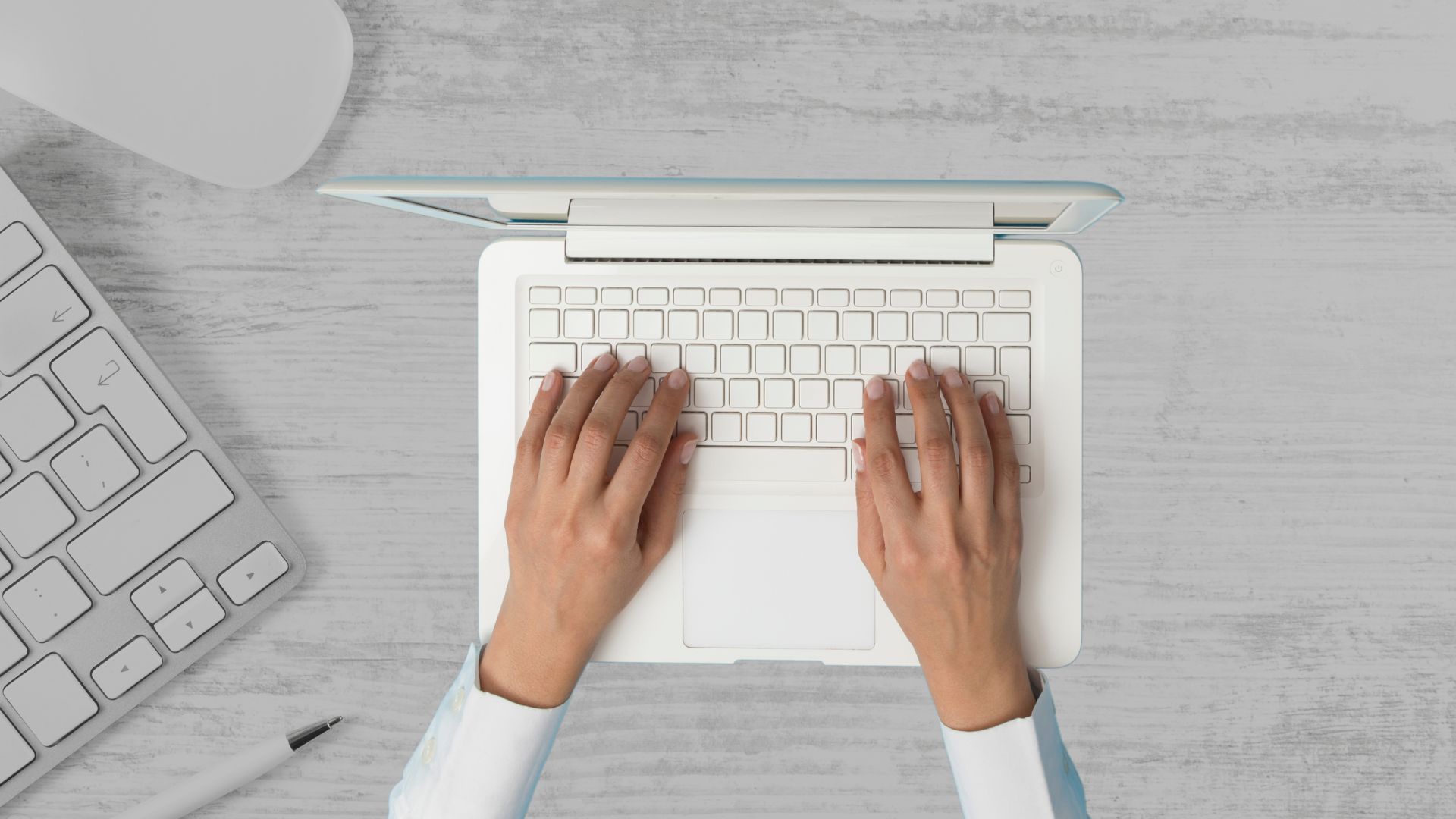 parallelstaff- A person types on a white laptop on a light wooden desk, with a wireless keyboard and a pen visible nearby.