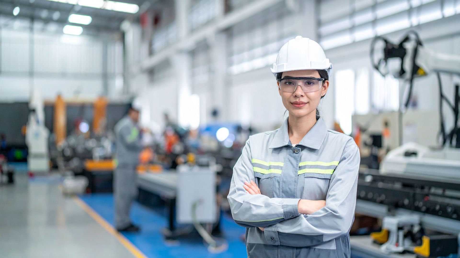 parallelstaff- A woman in safety gear, including a white hard hat and goggles, stands confidently with arms crossed in a modern industrial factory, with machinery and another worker visible in the background.