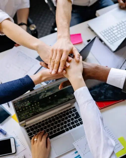 parallelstaff- A group of people stack their hands together over a desk with laptops, papers, and office supplies, symbolizing teamwork and collaboration in a work environment.