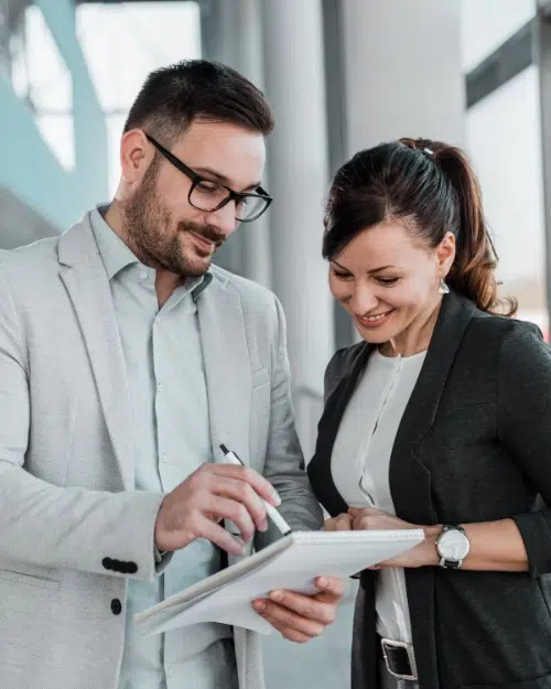 parallelstaff- Two professionally dressed people are standing indoors, smiling and looking at a notebook together. One is holding the notebook and a pen, while the other points at the page. They appear to be having a pleasant discussion.