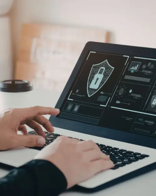 parallelstaff- A person types on a laptop displaying a cybersecurity interface with a large padlock icon and data charts, suggesting a focus on digital security. A coffee cup and blurred background are visible.