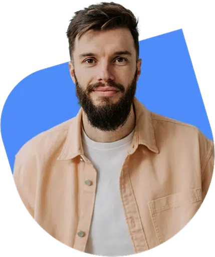 parallelstaff- A man with short brown hair and a full beard, wearing a beige button-up shirt over a white t-shirt, stands in front of a blue abstract background, smiling softly at the camera.