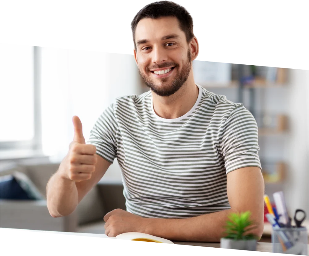 parallelstaff- A man with a beard, wearing a striped t-shirt, sits at a desk and smiles while giving a thumbs-up gesture. There are office supplies and a small plant on the desk in front of him.
