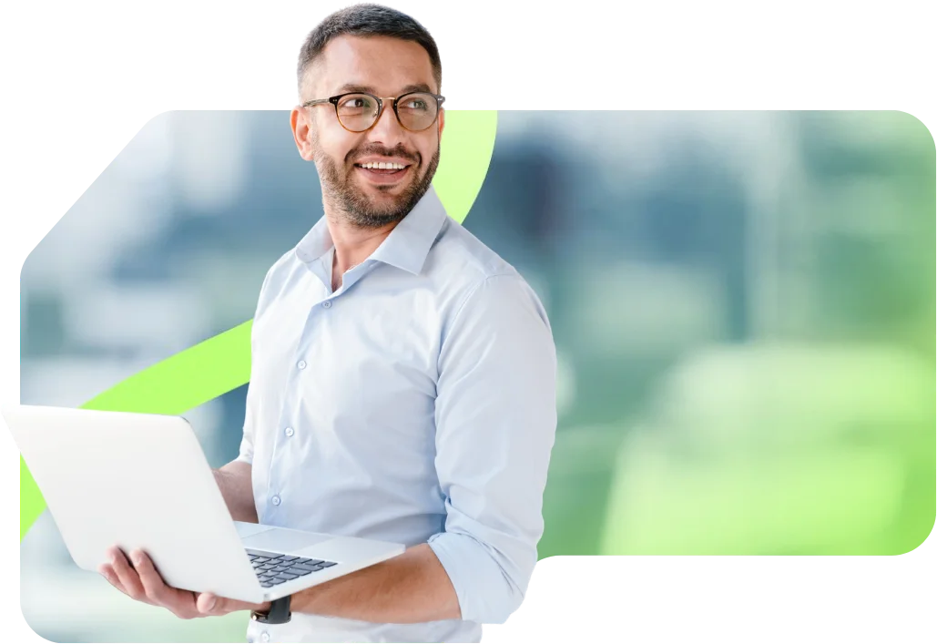 parallelstaff- A man wearing glasses and a light blue shirt smiles while holding an open laptop, standing in front of a blurred background with a green wavy line.
