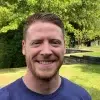 parallelstaff- A man with short red hair and a beard is smiling outdoors. He is wearing a blue shirt, and there are green trees and grass in the background on a sunny day.