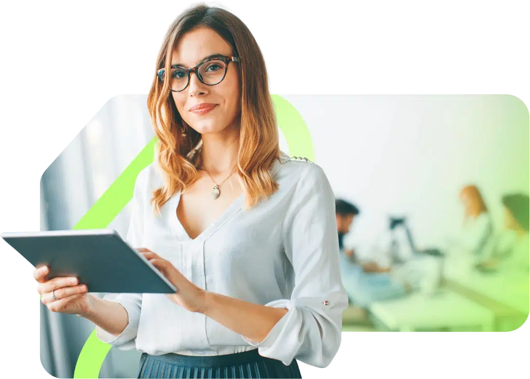 parallelstaff- A woman with glasses and shoulder-length hair is smiling while using a tablet. She is standing in an office environment with two blurred colleagues working at desks in the background.