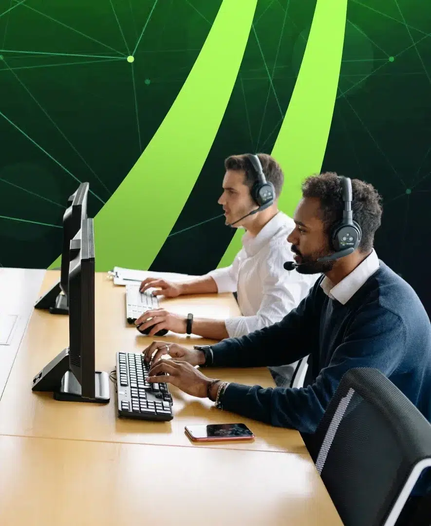 parallelstaff- Two men wearing headsets sit at a desk working on desktop computers, appearing focused. A smartphone is on the desk. The background features green and black digital abstract graphics.