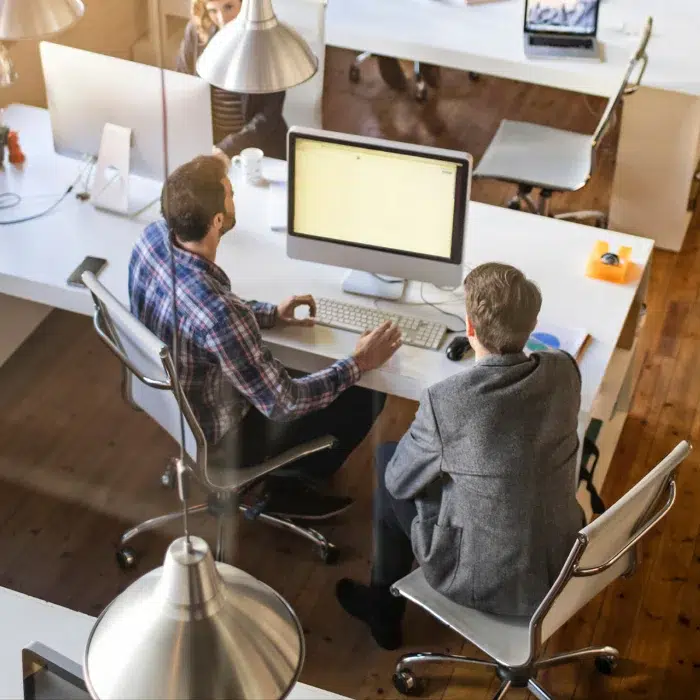 parallelstaff- Two people sit side by side at a desk, looking at a large computer monitor in a bright, modern office space with wooden floors and white desks. Other coworkers and office supplies are visible in the background.