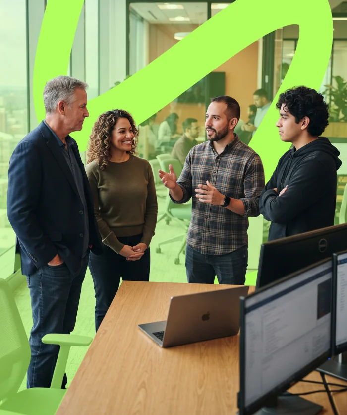 parallelstaff- Four people stand talking in an office by a wooden desk with two monitors and a laptop. Green designs overlay the image, and other people can be seen through a glass wall in the background.