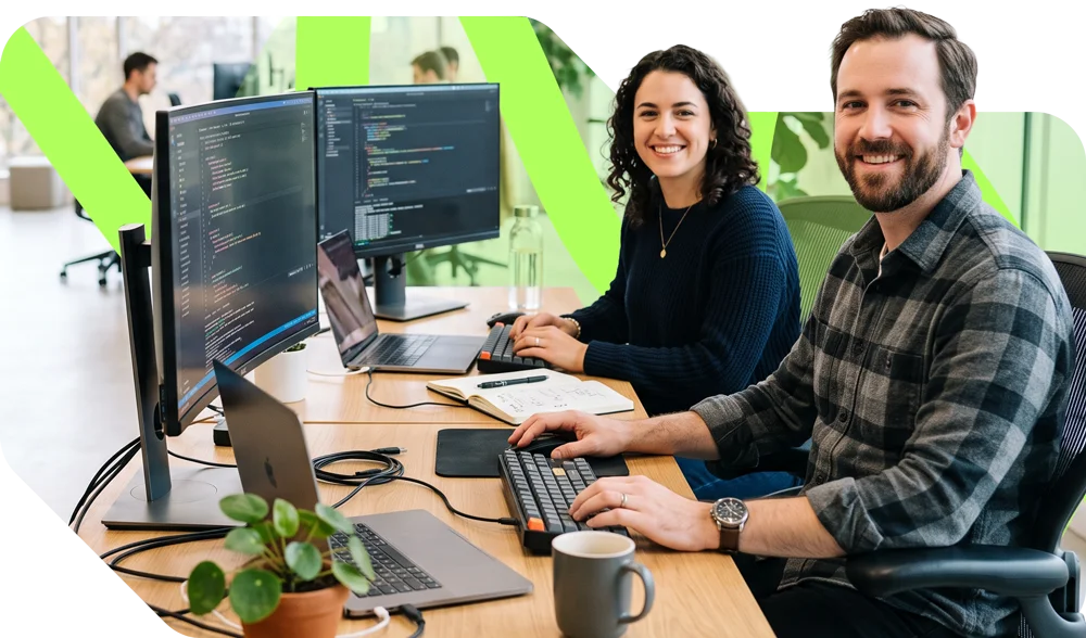 parallelstaff- Two people sit at a desk with computers, smiling at the camera. One types on a keyboard, while the other writes in a notebook. They appear to be working in a modern office space.
