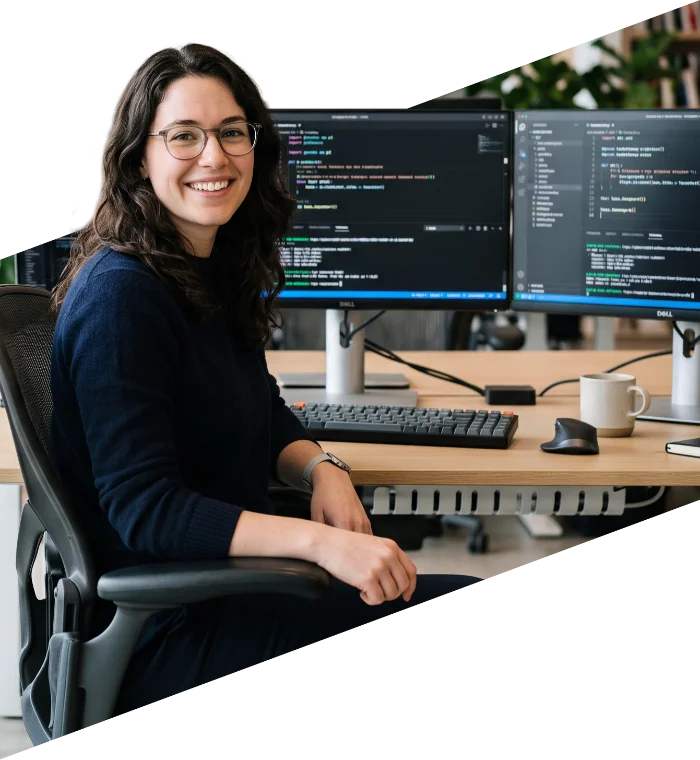 parallelstaff- A woman with glasses smiles while sitting at a desk with two monitors displaying code, a keyboard, mouse, and a coffee mug.