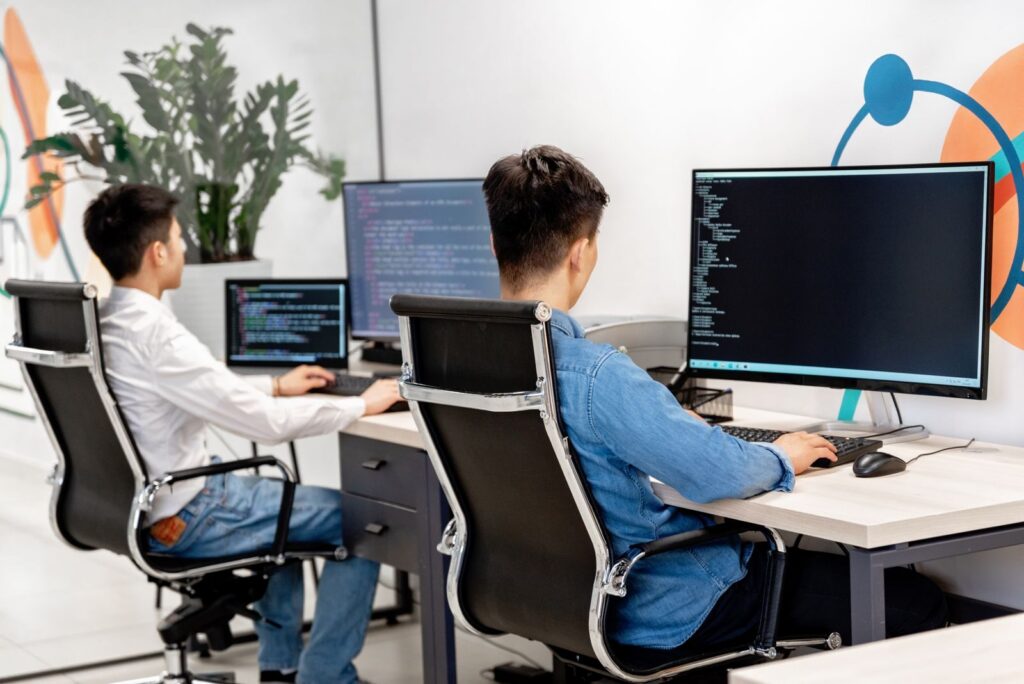 Two male developers at desks programming in a modern office workspace with large monitors.